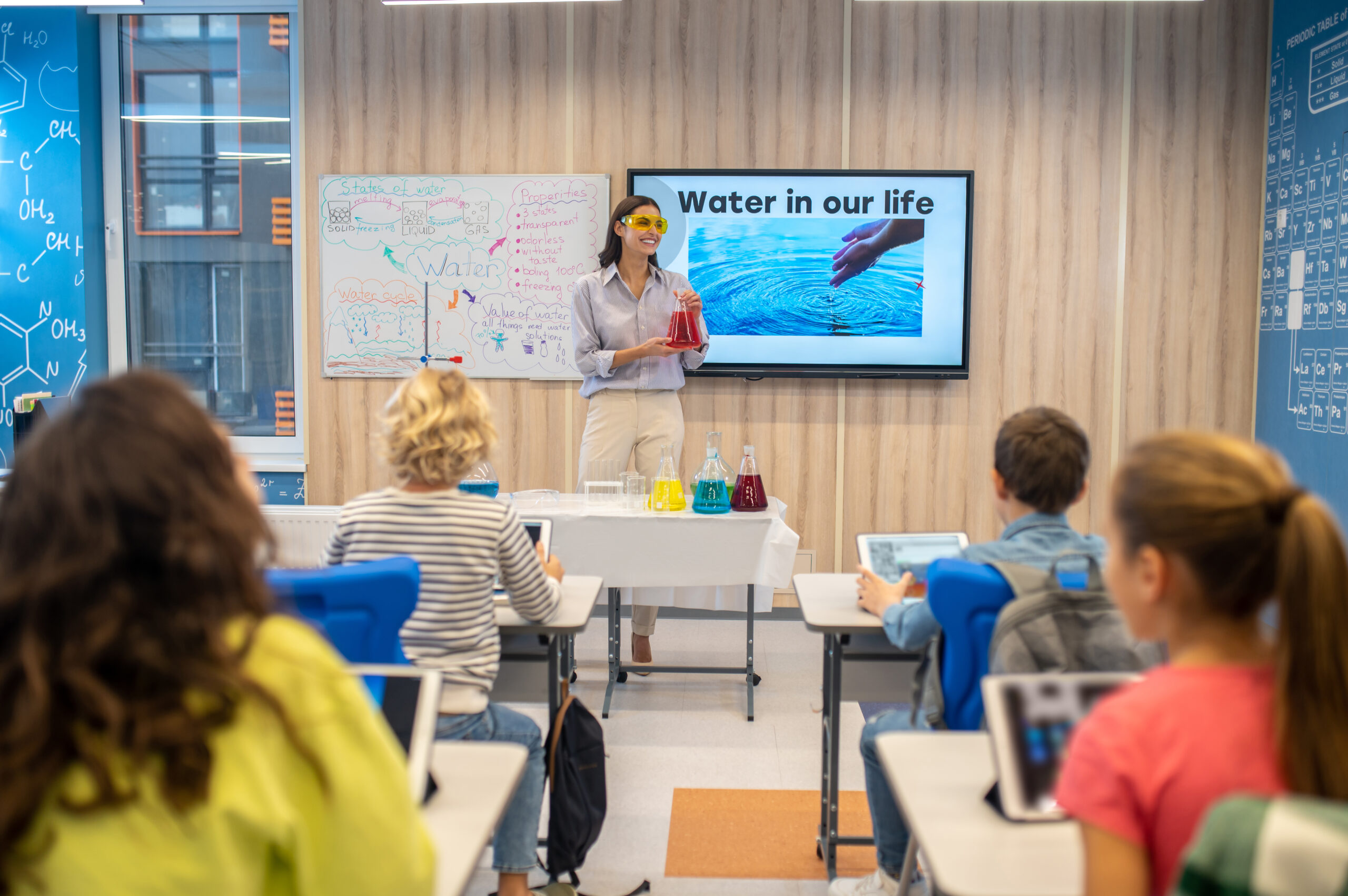 Chemistry lesson. Smiling woman in protective goggles holding flask with colored liquid standing looking at children in class